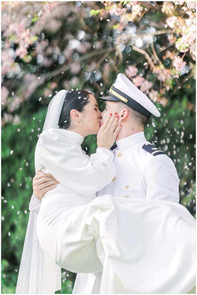 Close-up of the groom kissing the bride while holding her in his arms under falling pink petals in Bishop’s Garden during a Washington National Cathedral wedding.
