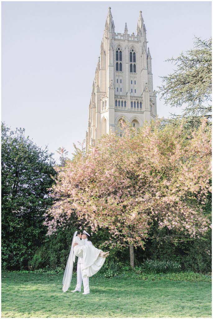 The groom carries the bride under a pink-blossomed tree in Bishop’s Garden during a Washington National Cathedral wedding, with the cathedral’s stone tower rising in the background.