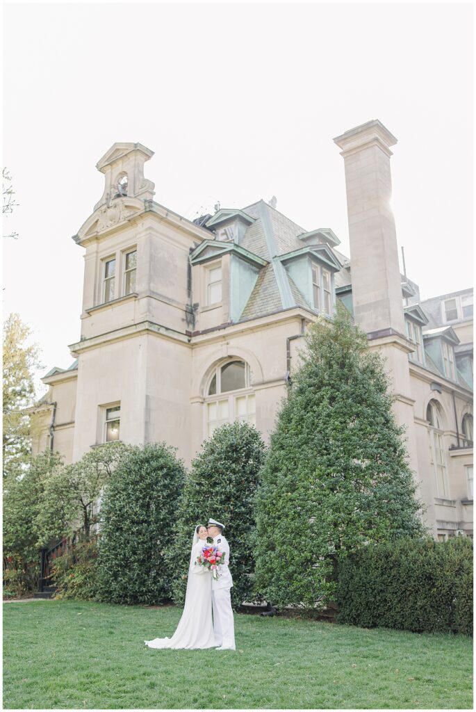 The bride and groom stand closely together on a lawn in front of a large stone building at the National Cathedral School, with the bride holding a colorful bouquet and the groom in a white naval uniform.