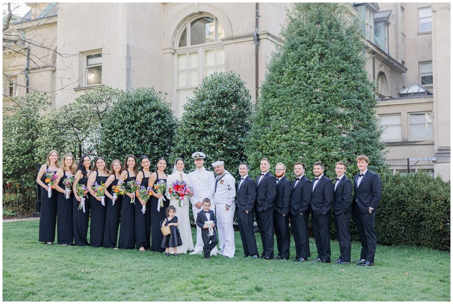 Full wedding party portrait at the National Cathedral School, with the bride and groom centered among bridesmaids in black dresses holding colorful bouquets and groomsmen in black tuxedos, accompanied by a young flower girl and ring bearer.