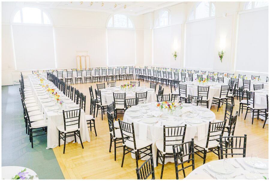 Wide view of a bright wedding reception room at the National Cathedral School, set with black chiavari chairs, round and long banquet tables covered in white linens, and small colorful floral arrangements.
