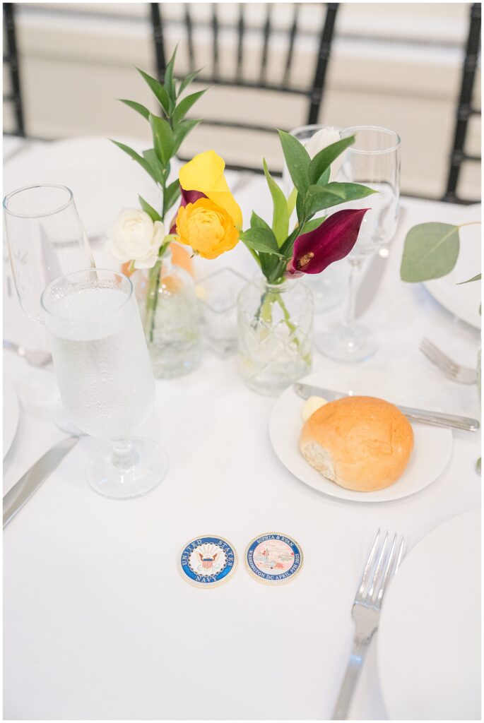 Close-up of a place setting at the National Cathedral School wedding reception, featuring a dinner roll, glassware, and two navy-themed challenge coins next to small bud vases with fresh flowers.