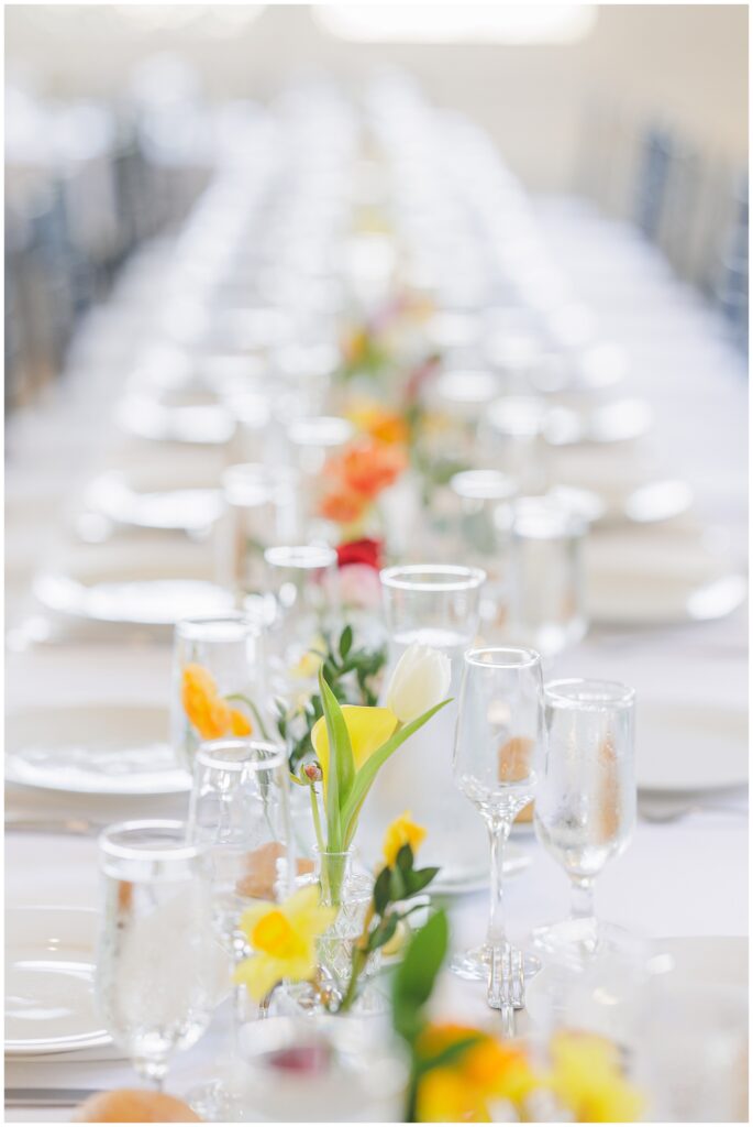 Focused view down the center of a long banquet table at the National Cathedral School wedding reception, showing rows of water glasses and bud vases filled with colorful spring flowers.