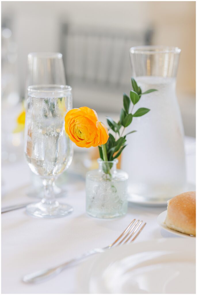 Detail shot of a table setting at the National Cathedral School wedding reception, featuring a single bright orange ranunculus in a small glass vase next to a water glass and bread roll.