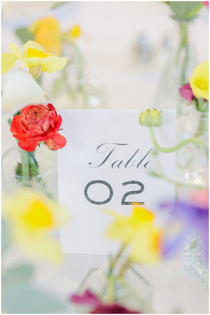 Table number card reading “Table 02” surrounded by an assortment of colorful spring flowers in clear bud vases at the National Cathedral School wedding reception.