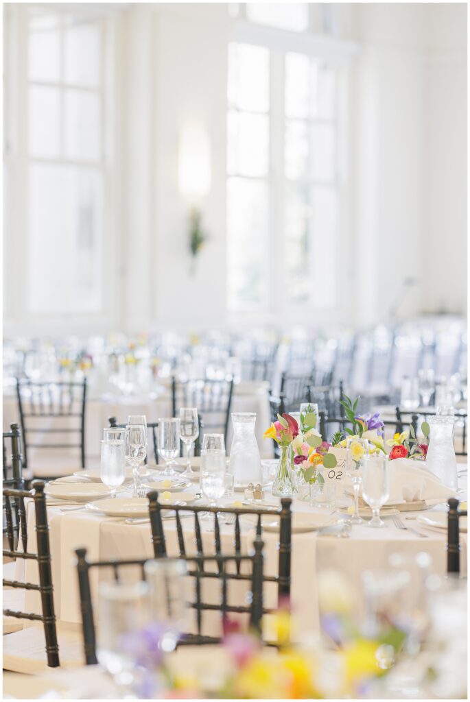 Overview of elegantly set round tables at the National Cathedral School wedding reception, decorated with floral centerpieces, clear glassware, and white linens, with large windows in the background.