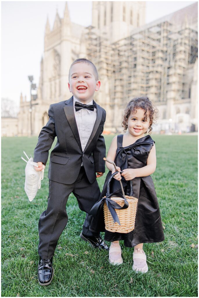 A young ring bearer in a black tuxedo and bow tie stands smiling next to a flower girl in a black satin dress holding a small basket, with the Washington National Cathedral in the background.
