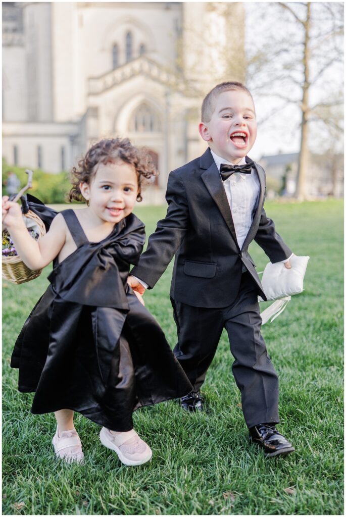 A joyful ring bearer and flower girl run hand-in-hand on the lawn outside the Washington National Cathedral, both dressed in black formalwear and smiling broadly.