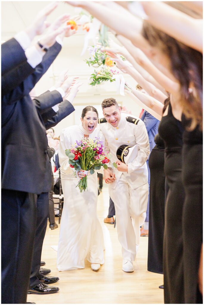 The bride and groom, dressed in a long-sleeved gown and a white naval uniform, joyfully walk through a tunnel of raised arms and flowers formed by their wedding party at a National Cathedral School wedding reception.