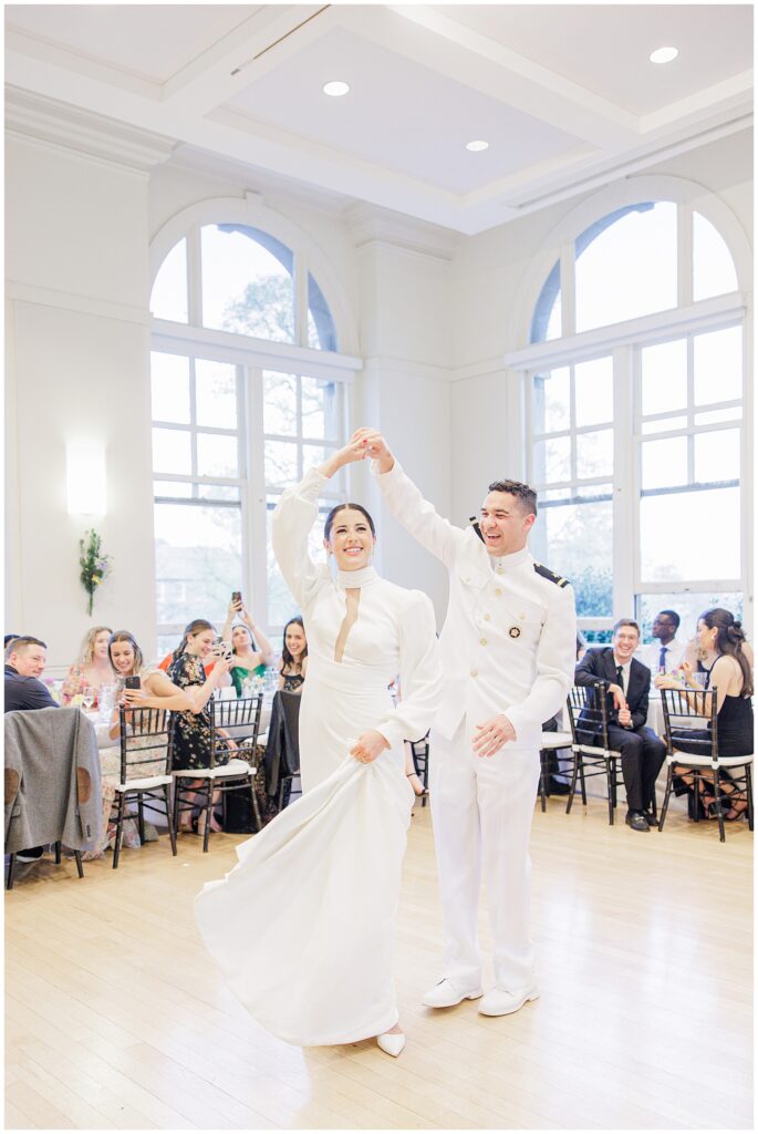 The bride and groom smile and twirl together during their first dance at a National Cathedral School wedding reception, surrounded by seated guests.