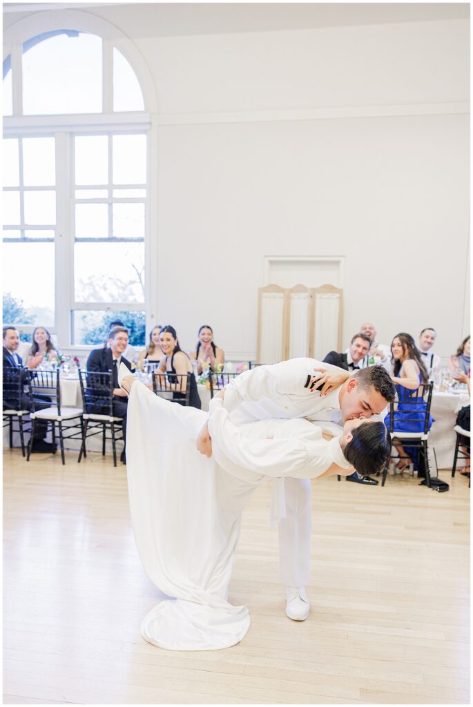 The groom dips the bride and kisses her during their first dance at a National Cathedral School wedding reception, with guests watching and smiling in the background.