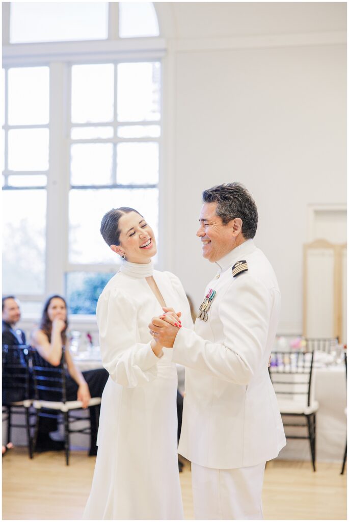 The bride shares a joyful dance with her father, both smiling, during a National Cathedral School wedding reception; he wears a white military uniform with medals.