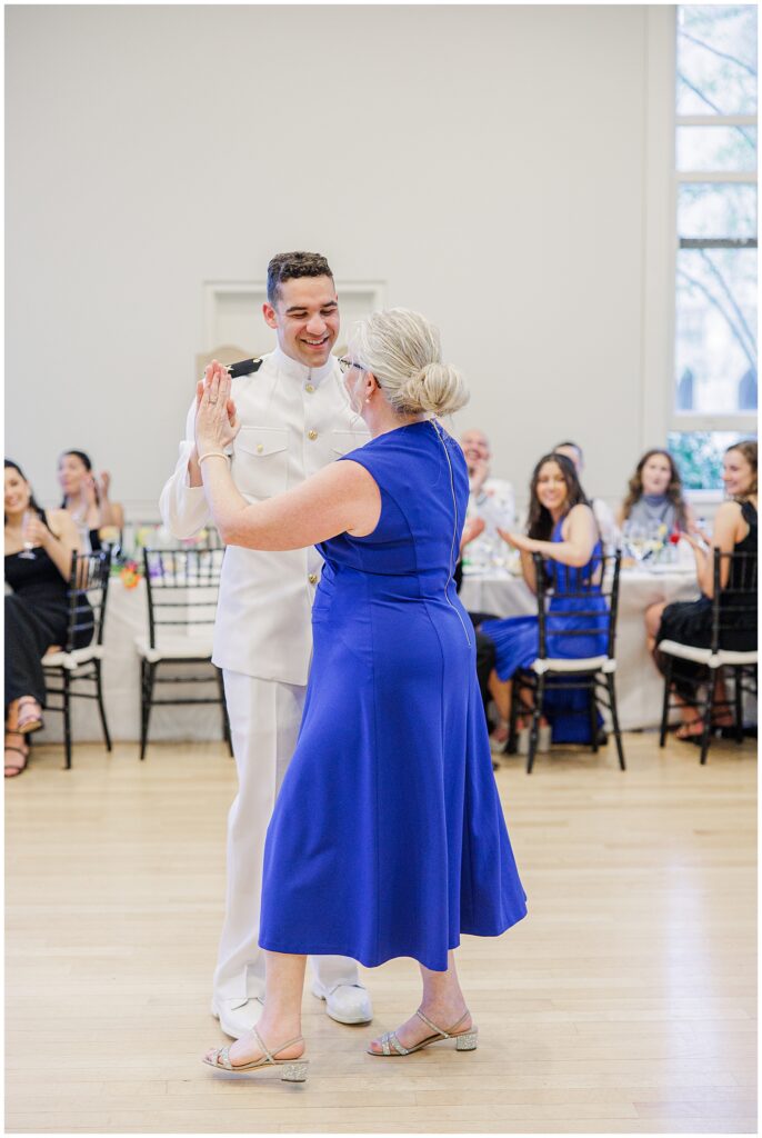 The groom dances with his mother, who wears a royal blue dress, during a National Cathedral School wedding reception as guests watch and applaud.