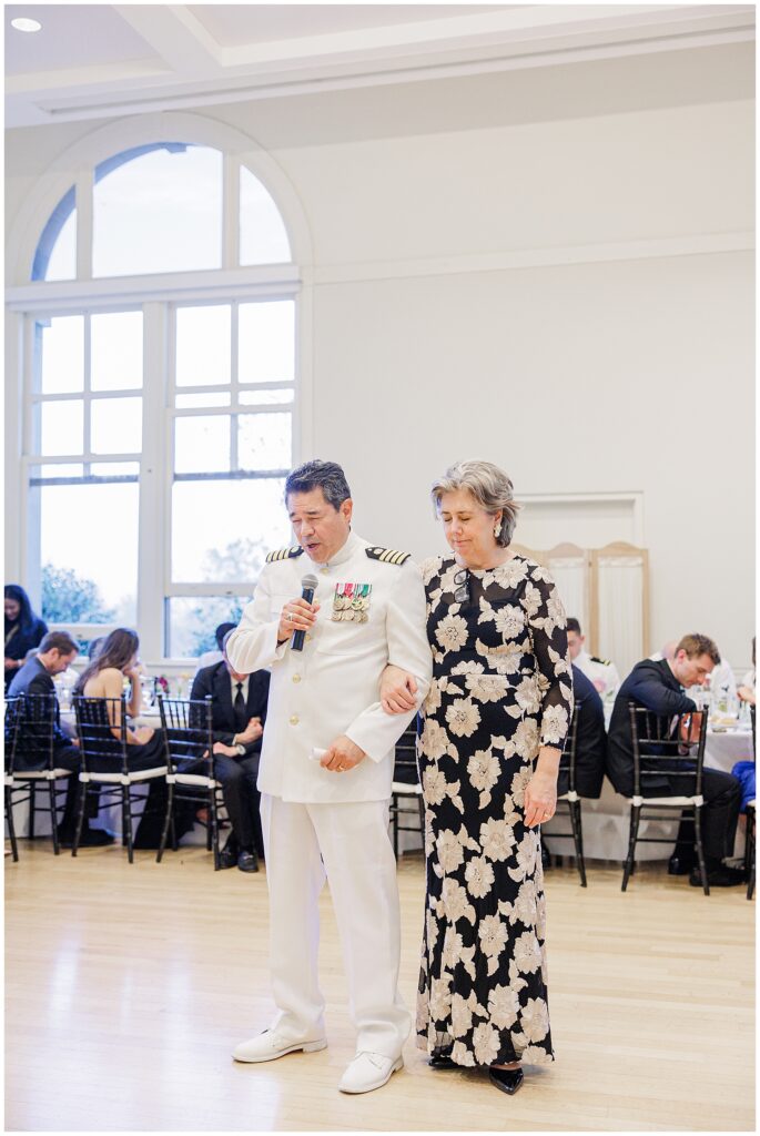 A man in a white military uniform with medals speaks into a microphone while linking arms with a woman in a floral dress during a National Cathedral School wedding reception.