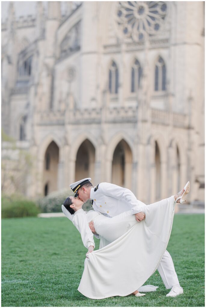 The groom dips and kisses the bride on the lawn in front of the Washington National Cathedral, with her dress flowing gracefully.