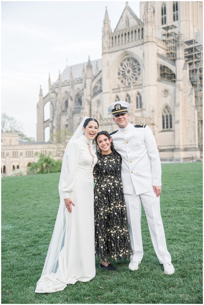 The bride and groom stand smiling with a guest in a black and gold dress on the lawn outside the Washington National Cathedral.
