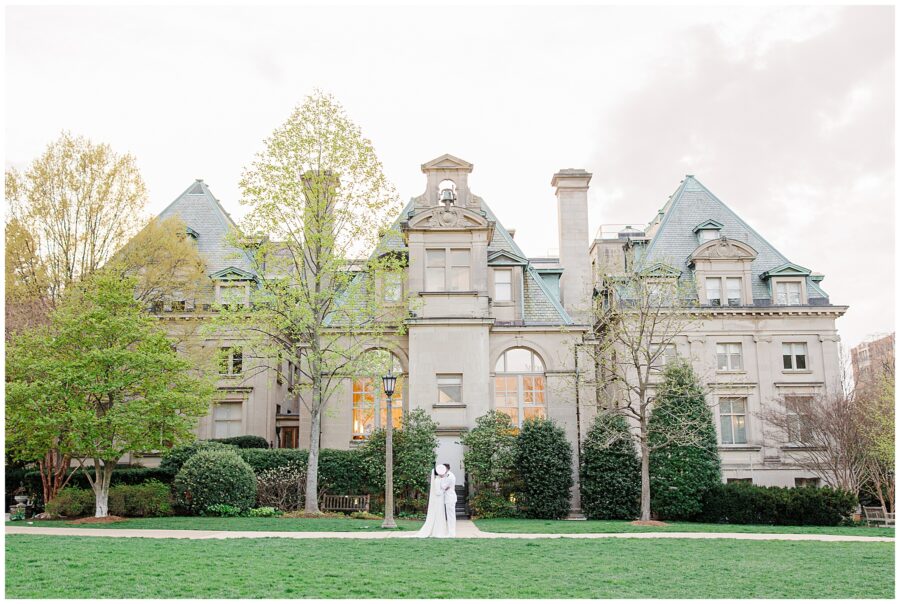 The bride and groom share a kiss in front of a grand stone building with dormer windows and green roofing at the Washington National Cathedral.