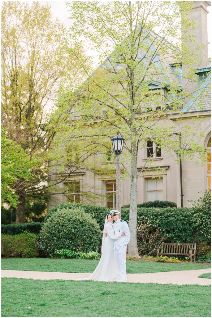 The bride and groom embrace near a vintage-style lamppost and tall spring trees in a garden area at the Washington National Cathedral.