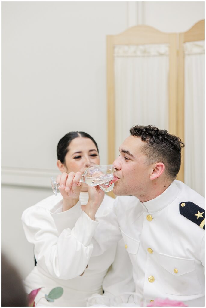 Bride and groom at a National Cathedral School wedding reception link arms and sip from champagne glasses while seated.