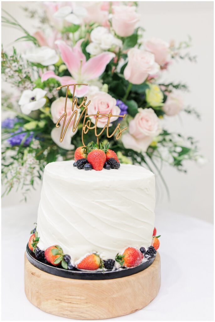 A white frosted wedding cake topped with berries and a wooden “The Motts” topper sits on a wooden stand, with a floral arrangement behind it.