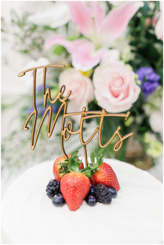 Close-up of the wedding cake topper reading “The Motts” placed among fresh strawberries and berries, with pink and purple flowers in the background.