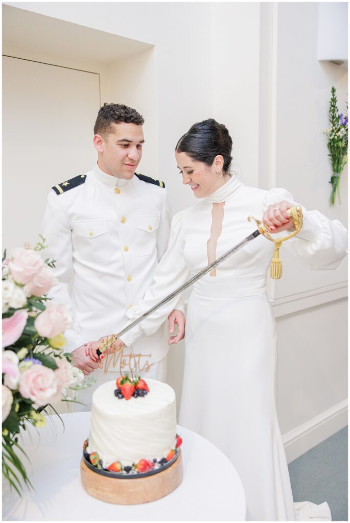 The bride and groom, dressed in white, cut their wedding cake with a ceremonial sword at their National Cathedral School reception.