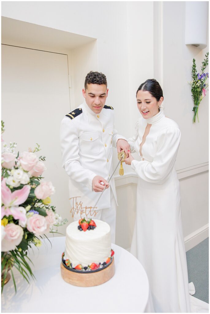 The couple begins slicing the wedding cake together using a sword, surrounded by floral decorations at their National Cathedral School reception.