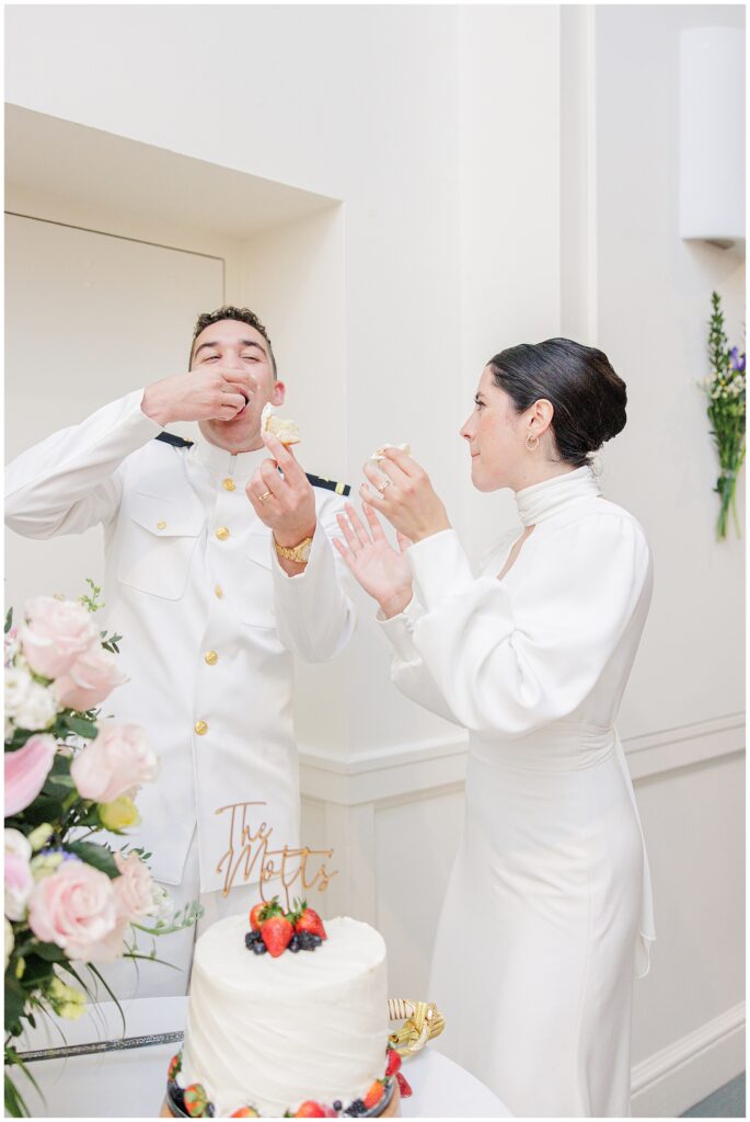 Groom enthusiastically eats cake while the bride looks on, both enjoying the cake-cutting moment at their National Cathedral School reception.