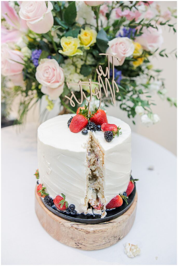 The wedding cake with a slice cut out, revealing a fruit-filled interior, sits on a table decorated with flowers and berries at the National Cathedral School reception.