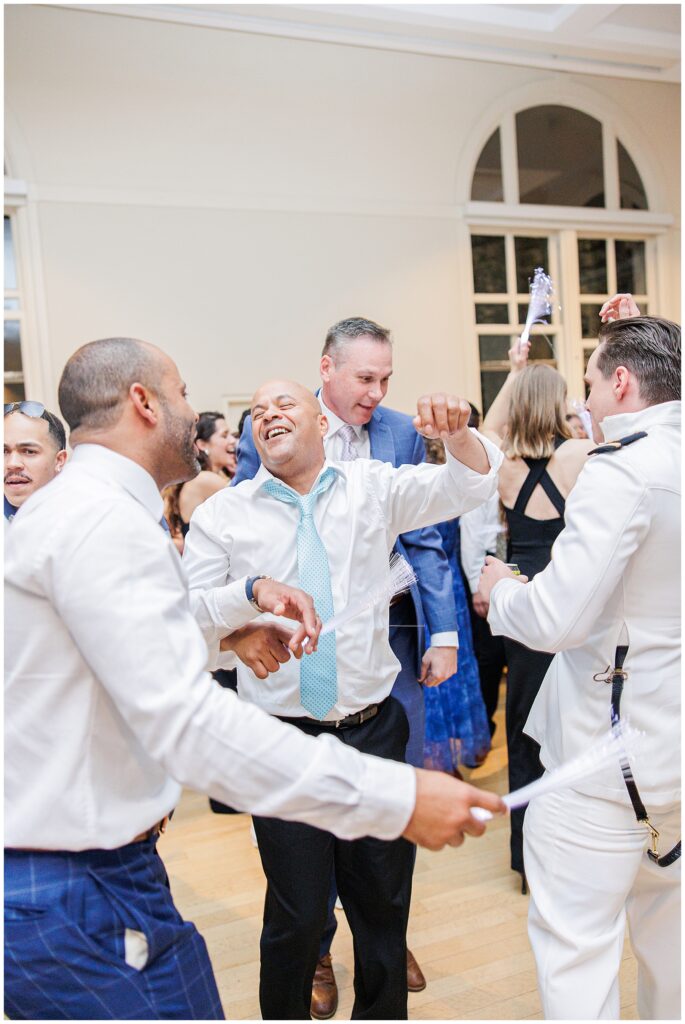 Guests joyfully dance together on the reception floor at the National Cathedral School, with arms raised and smiles all around.