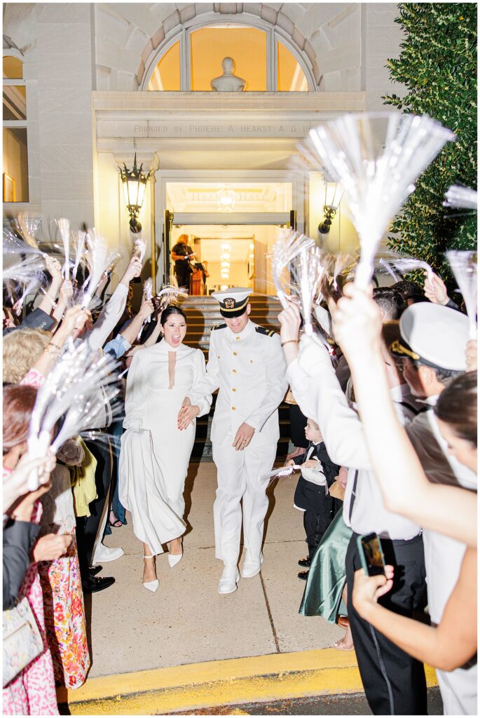 The bride and groom walk hand-in-hand, smiling, as guests cheer and wave light-up streamers during their exit from the National Cathedral School reception.