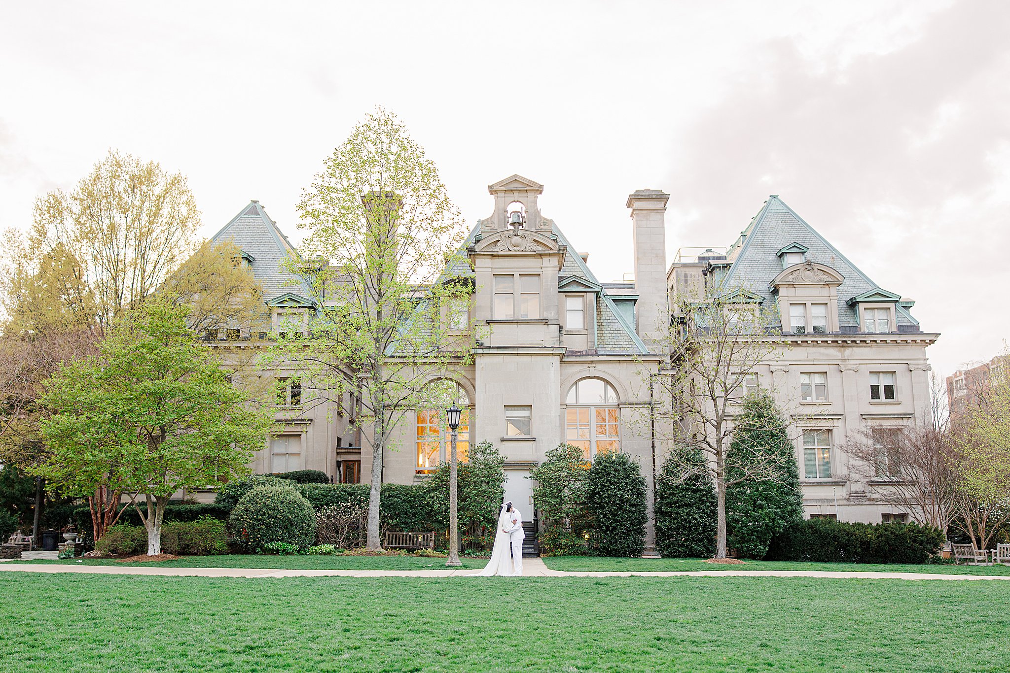 The bride and groom stand closely together on a green lawn in front of Hearst Hall at the National Cathedral School during golden hour.