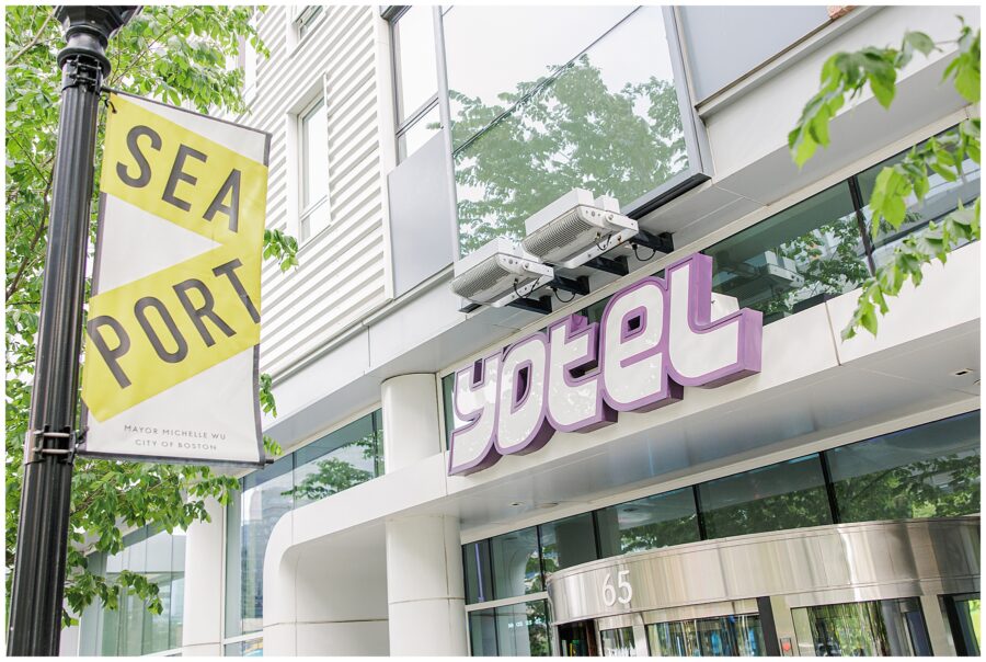 Exterior of the YOTEL hotel in Boston’s Seaport district, with a purple and white “yotel” sign and a nearby street banner reading “SEA PORT” on a yellow and white background.