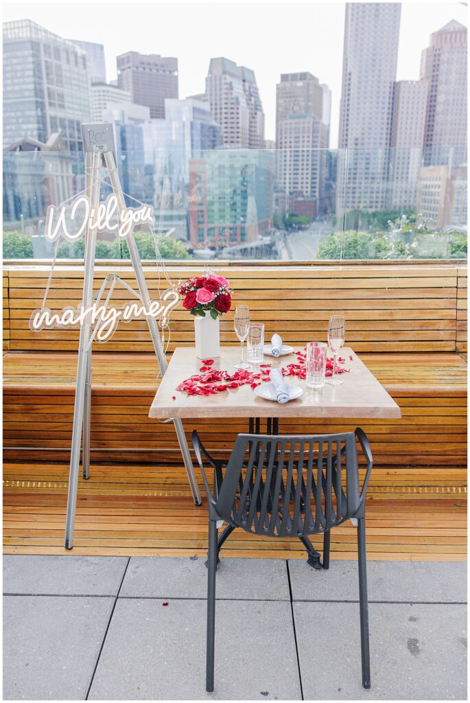 A rooftop table set for a proposal, with a vase of red and pink roses, rose petals scattered on the table, champagne glasses, and a neon sign that reads “Will you marry me?” The Boston skyline is visible in the background.