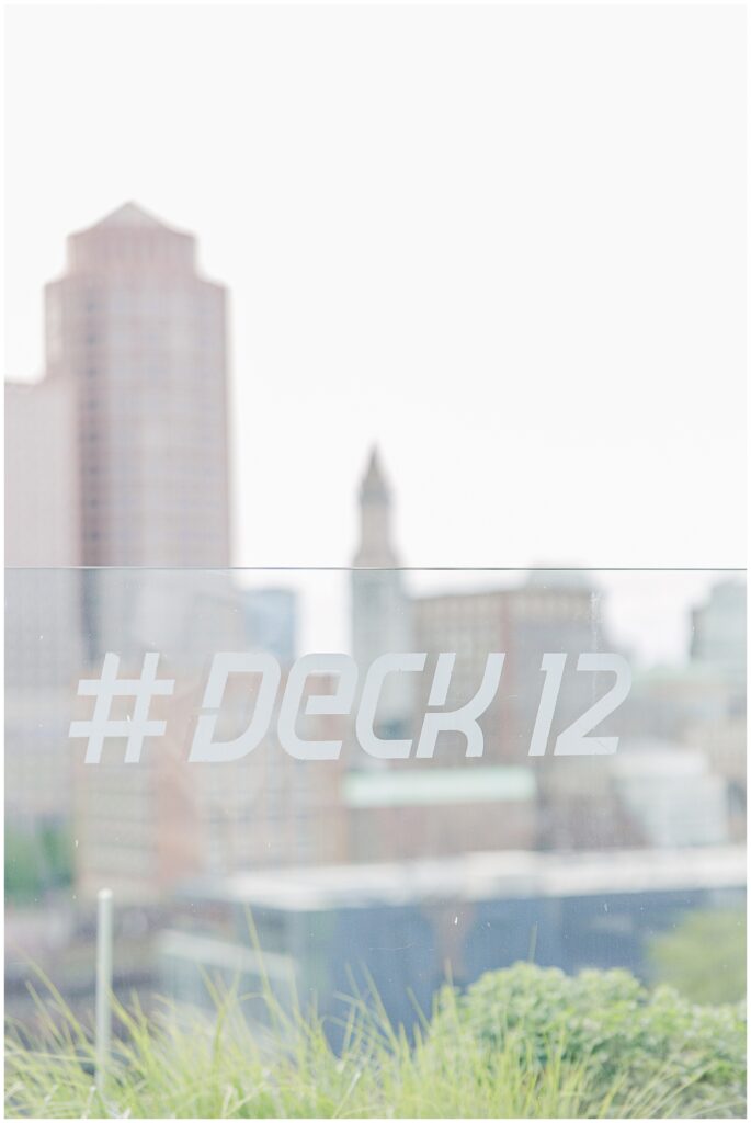 Close-up of a glass barrier on the rooftop with “#DECK 12” etched on it. The blurred Boston skyline is in the background.