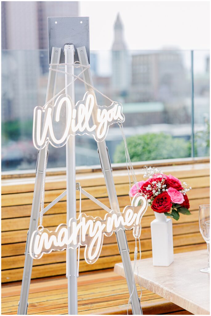 Close-up of the “Will you marry me?” neon sign hanging on a tripod on the rooftop, with a vase of red and pink roses in the background.