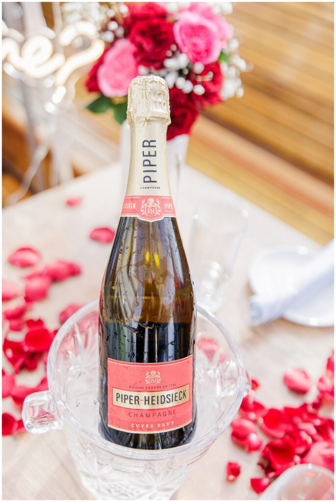 Close-up of a bottle of Piper-Heidsieck champagne in an ice bucket, placed on a rooftop table decorated with rose petals and set for the proposal.