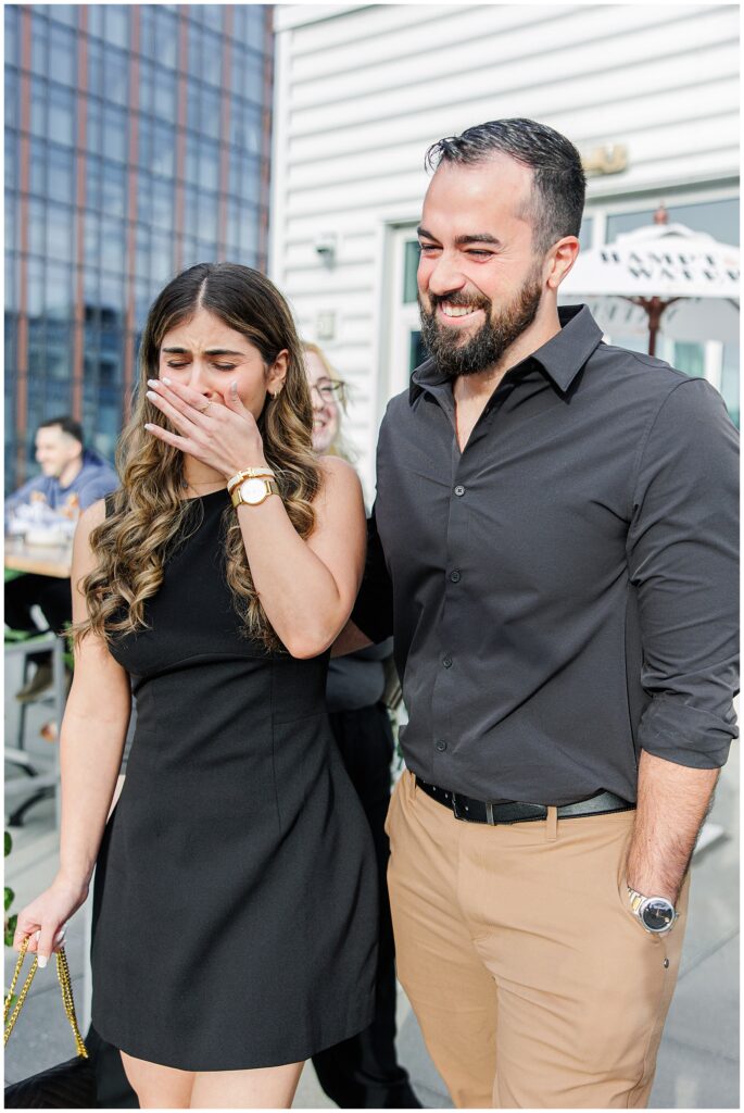 A woman in a black dress looks emotional and covers her mouth with her hand, standing next to a smiling man in a black shirt and tan pants on the rooftop at Deck 12.