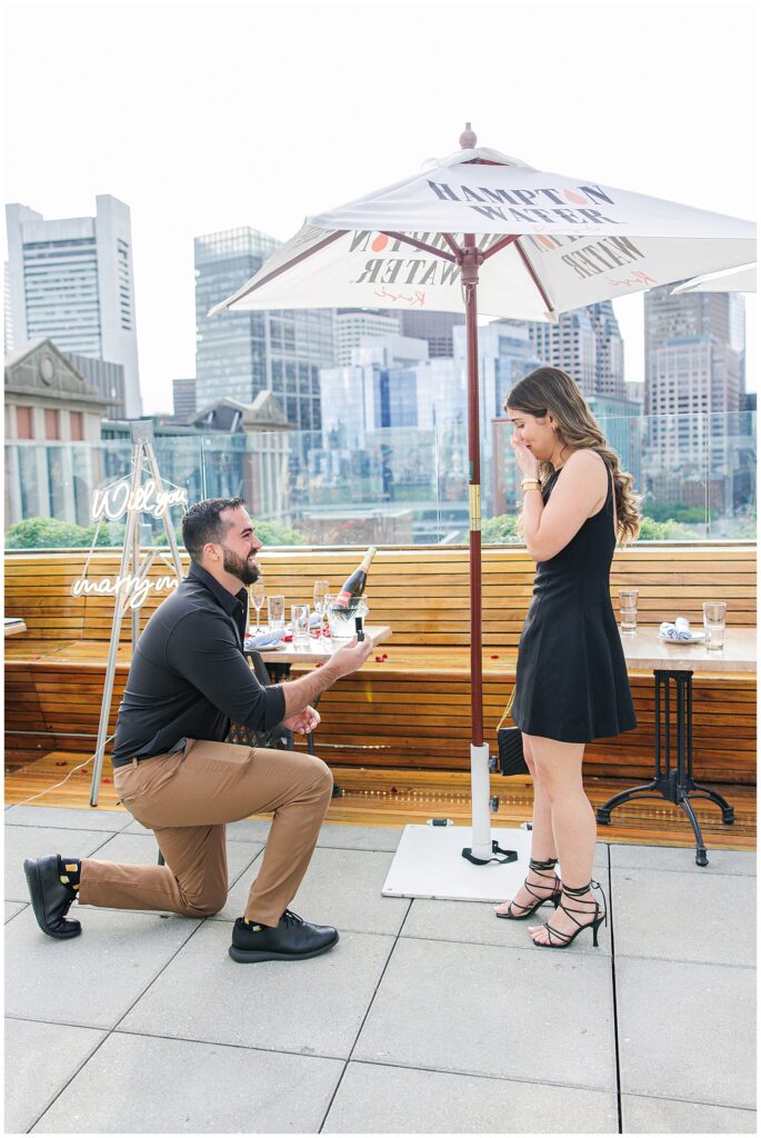 A man kneels on one knee, holding an engagement ring box toward a surprised woman standing in front of him. A “Will you marry me?” neon sign and champagne are on a table behind them, with the Boston skyline in the background.