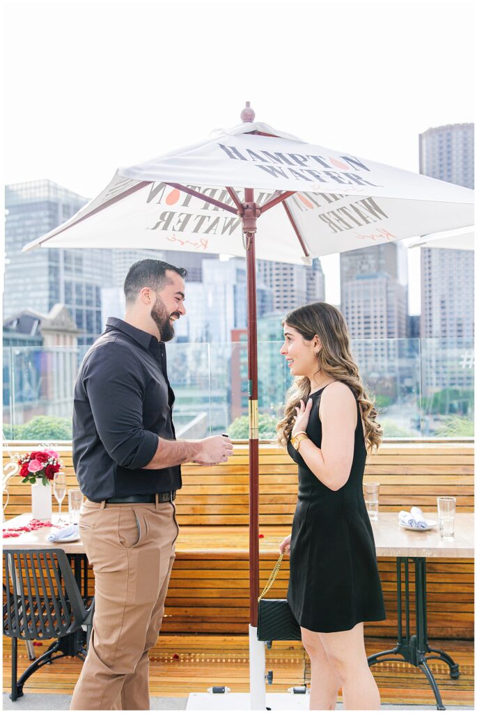 The couple stands under a white umbrella on the rooftop. The man is holding an engagement ring, and the woman reacts emotionally with a hand to her chest.