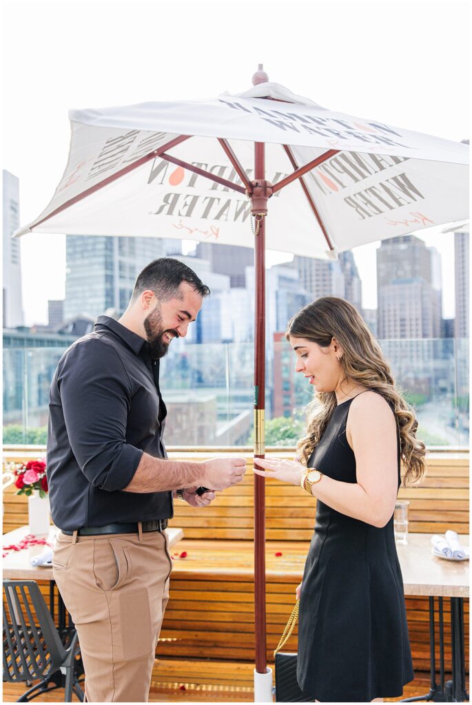 The man places an engagement ring on the woman’s finger. Both are smiling and standing on the rooftop under a white umbrella, with the Boston skyline behind them during their Deck 12 Boston proposal.