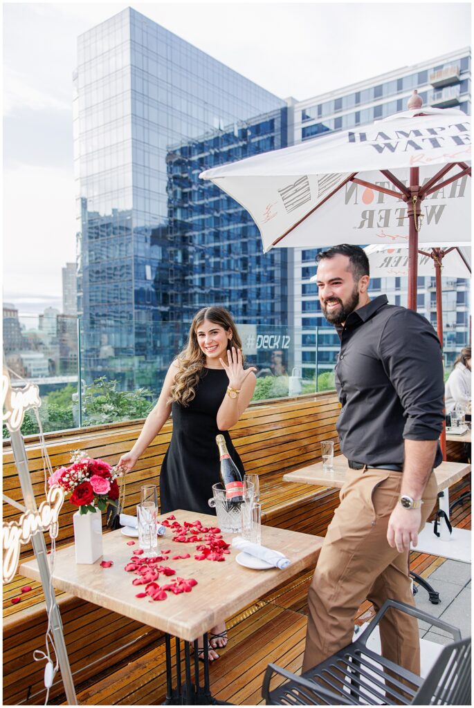 Woman in a black dress smiling and showing her engagement ring while standing next to a man on the rooftop at Deck 12 Boston. The table is decorated with rose petals, champagne, and flowers, with the Boston skyline in the background.