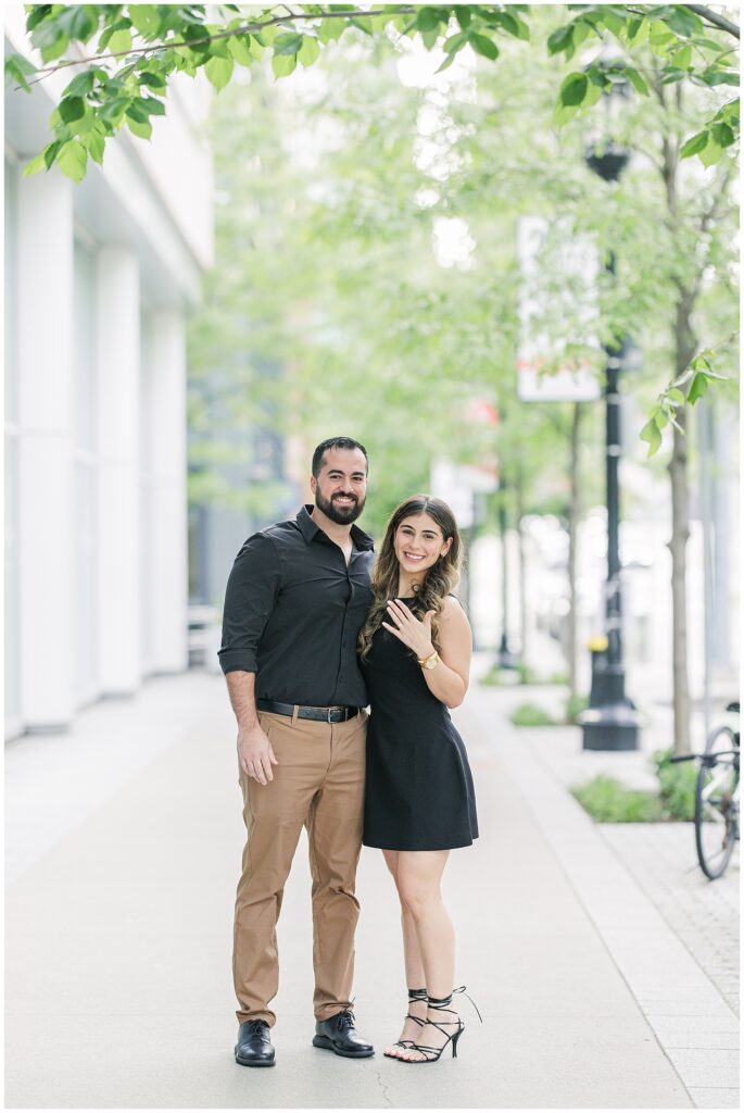 Couple standing on a tree-lined sidewalk near the Seaport in Boston. The woman smiles and shows off her engagement ring, while the man stands with his arm around her.