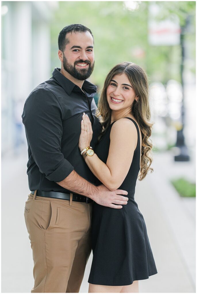 Close-up portrait of the couple standing on the sidewalk. Both are smiling and embracing, with the woman’s engagement ring visible.