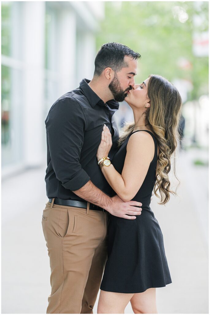 The couple shares a kiss on the sidewalk. The woman’s hand with her engagement ring is visible on the man’s chest.