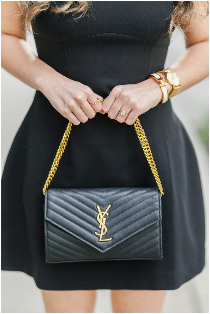 Close-up of the woman holding a black Yves Saint Laurent handbag with a gold chain strap. A diamond engagement ring is visible on her finger, along with a gold watch and bracelet.
