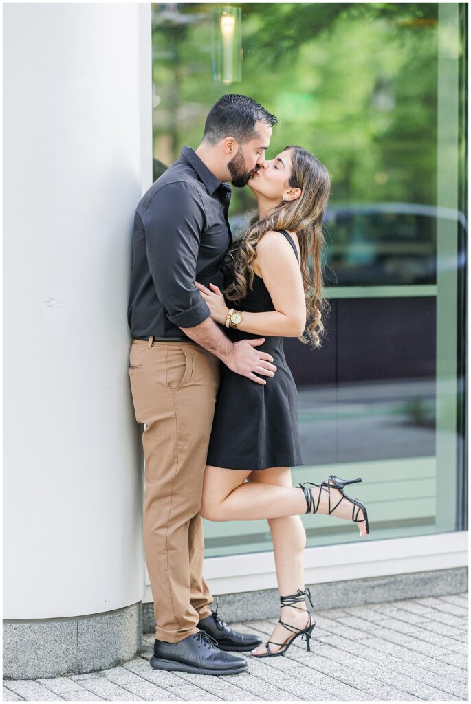The couple kisses while standing against a white column outside, with the woman’s leg bent behind her. The engagement ring is visible on her left hand.