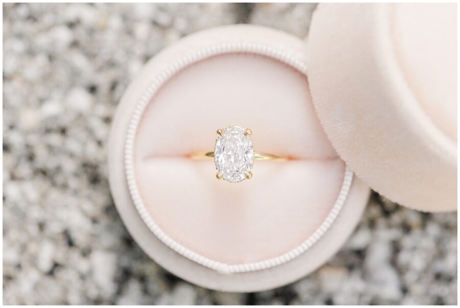 Close-up of a diamond engagement ring with an oval-cut stone set in a gold band, displayed in a round pink velvet ring box.