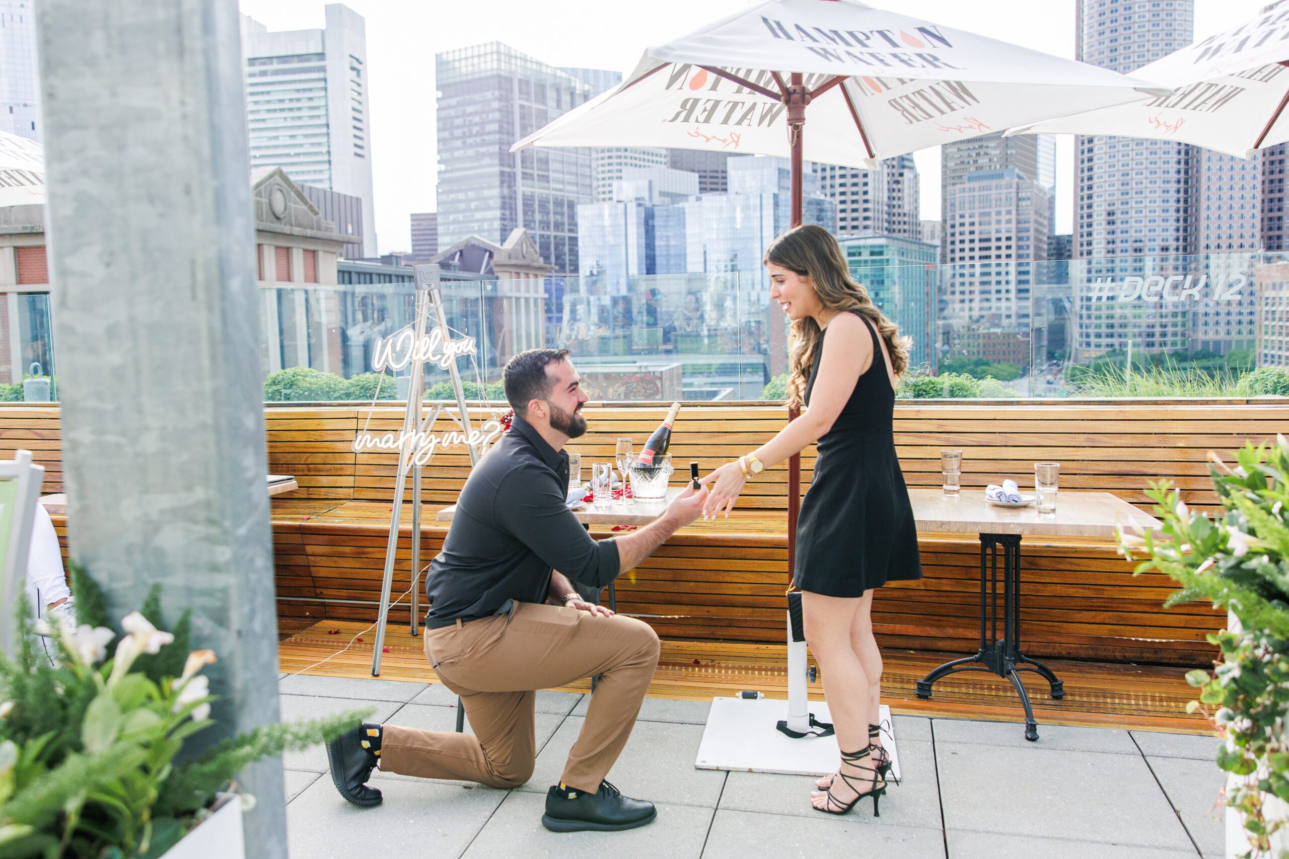 Man kneeling and holding the woman’s hand during a Deck 12 Boston proposal on the rooftop. The woman looks excited, and the table behind them is decorated with champagne and rose petals, with the Boston skyline in the background.