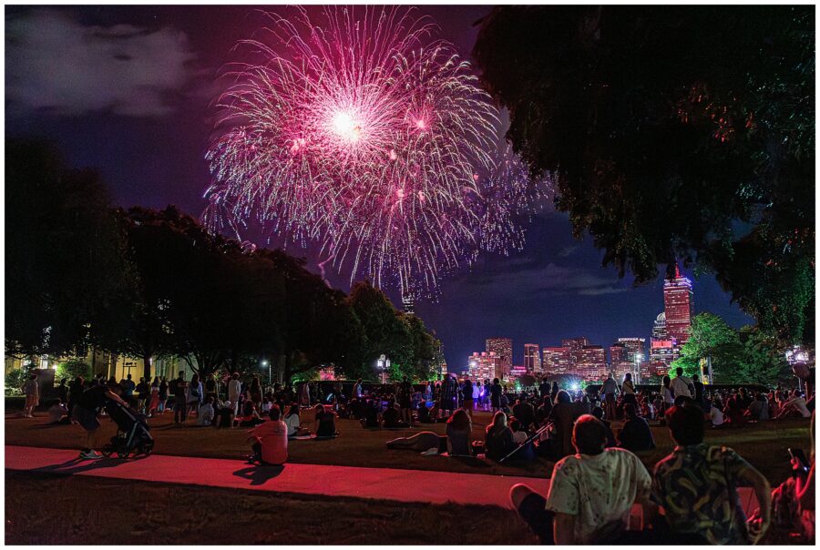 People gathered on a lawn watching vibrant fireworks over the Boston skyline during a 4th of July celebration.